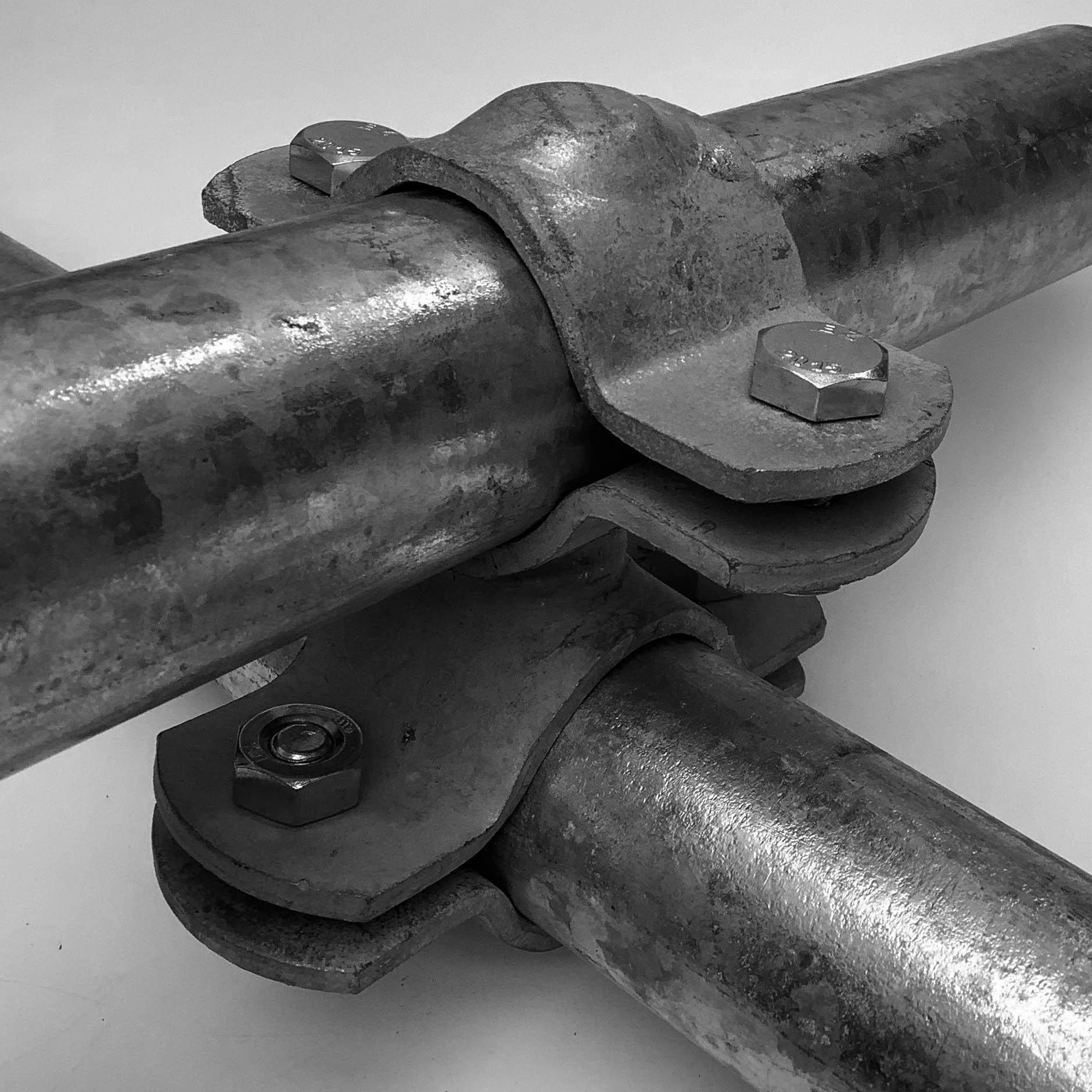 Grauer, robuster Überkreuz-Verbinder aus Metall verbindet zwei Rohre im 90°-Winkel; das obere verläuft horizontal über dem unteren. Mit vier M10x35 Schrauben fixiert, umschließt er 101,6 mm und 48,3 mm Rohre. Oberfläche leicht rau, reflektierend.