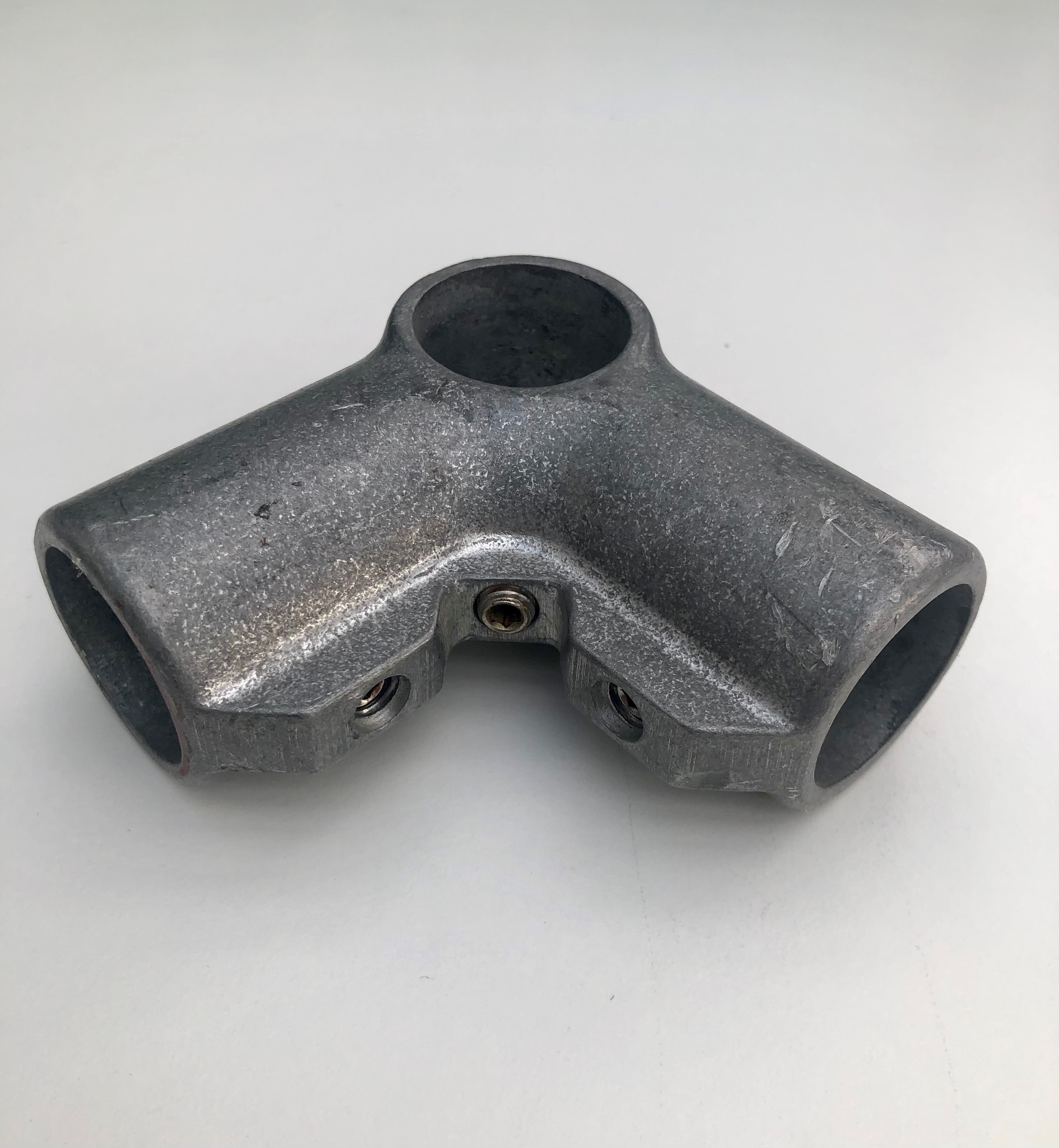 Grauer Eckverbinder aus rohem Aluminium, verbindet drei Rohre im 90-Grad-Winkel. Mit drei runden Öffnungen zum Einführen der Rohre und drei sichtbaren Madenschrauben zur sicheren Fixierung. Oberfläche leicht rau, unbehandelt.