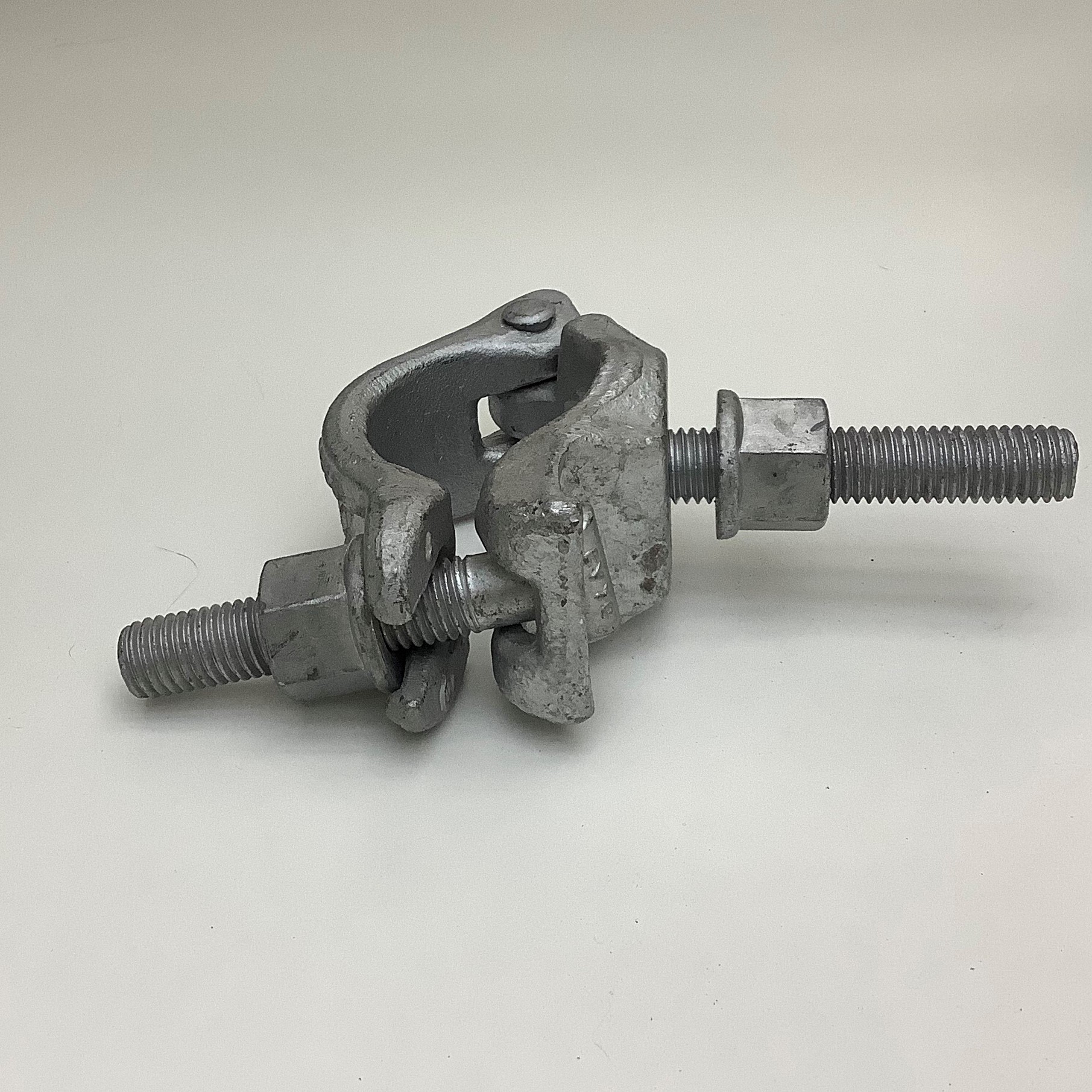 Ein robuster, grauer Metallgelenkverbinder, vermutlich eine Gerüstklemme, liegt auf weißem Hintergrund. Er besteht aus zwei groben, scharnierten Metallteilen, gehalten von zwei Gewindebolzen mit Muttern. Rau, industriell, leicht abgenutzt.
