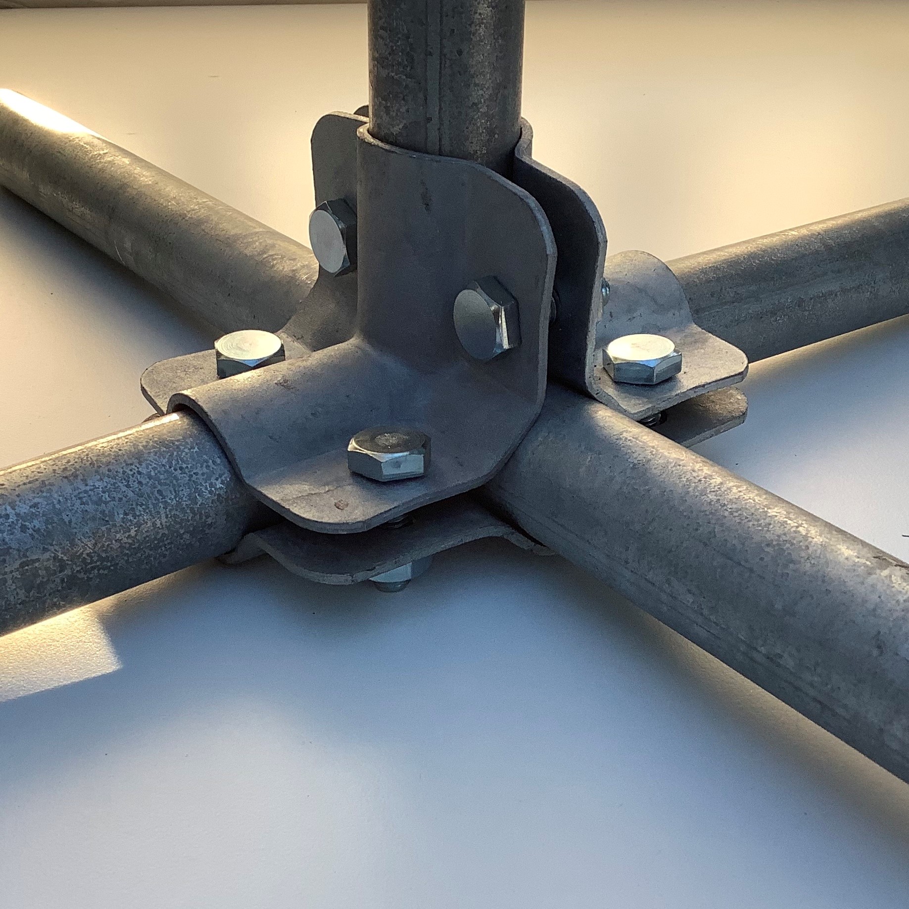 Conector en cruz de metal galvanizado con cuatro tubos horizontales en ángulo de 90 grados. Un tubo vertical atraviesa el centro de la sólida unión por presión. Varios tornillos hexagonales plateados fijan los tubos. Acabado metálico rugoso.