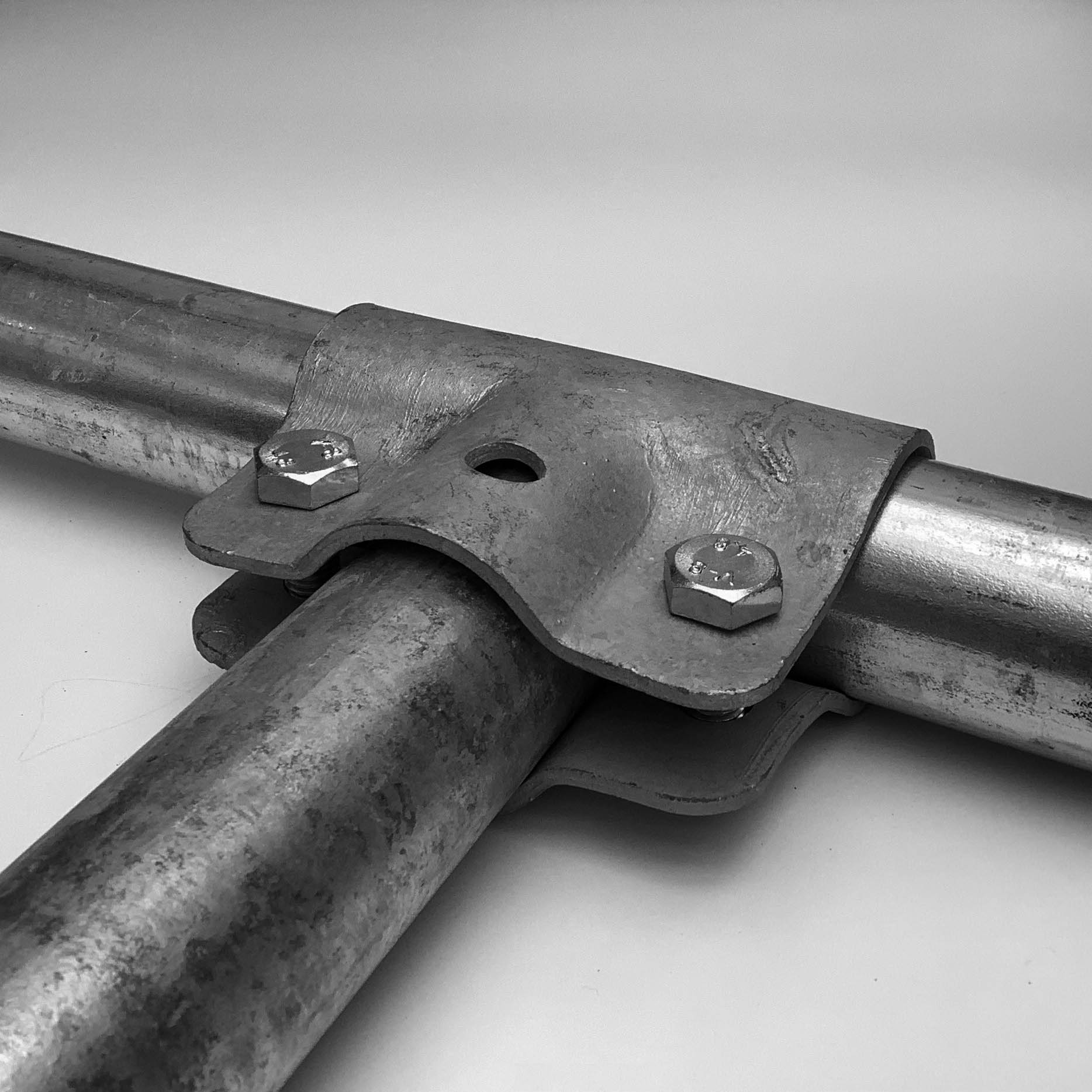 Ein verzinkter T-Verbinder, der zwei Metallrohre im 90-Grad-Winkel miteinander verbindet. Zwei Schrauben mit Sechskantköpfen fixieren die Rohre fest in der langen Klemmung. Die Oberfläche von Verbinder und Rohren zeigt eine raue, metallische Textur.