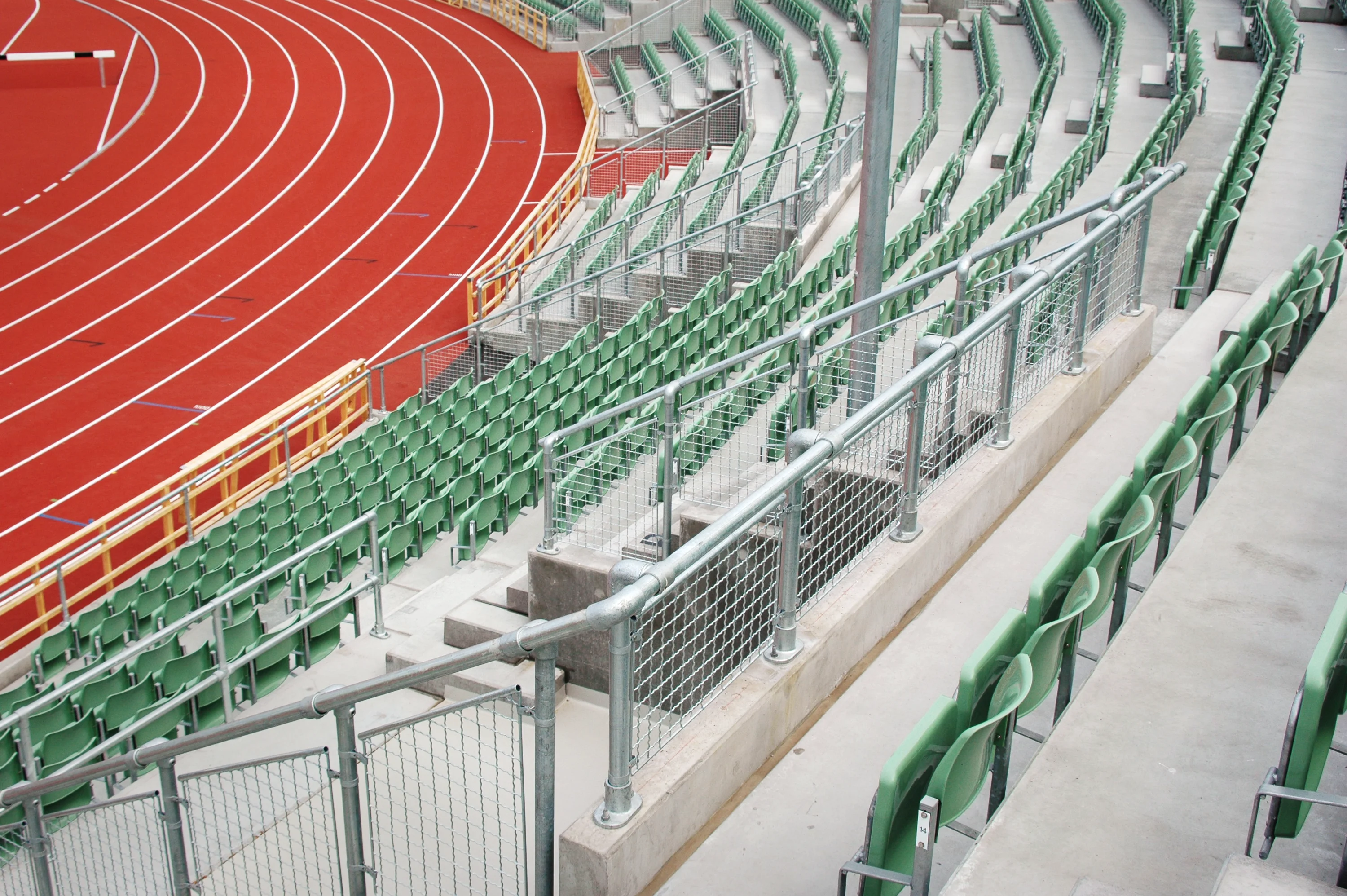 A la izquierda hay una pista de atletismo roja y vacía con líneas blancas de carril que gira a la derecha en la curva. A la derecha y al fondo se ven gradas verdes y vacías sobre escalones de hormigón, separadas por barandillas metálicas con rejillas.