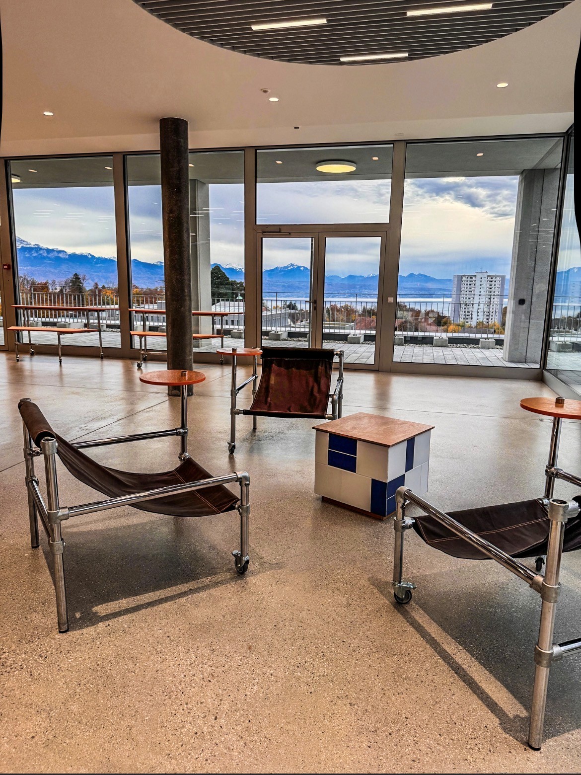 Moderna zona de salón con grandes ventanales y amplias vistas al lago, a las montañas nevadas y al cielo nublado. Tres sillones de cuero marrón con estructura cromada y ruedas, así como pequeñas mesas naranjas, descansan sobre un suelo de terrazo claro.